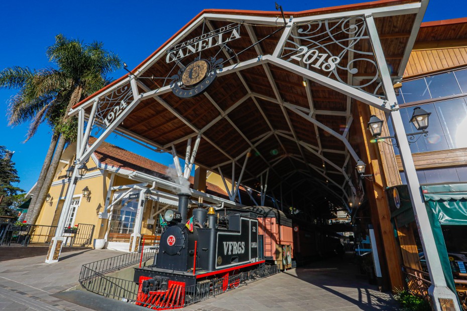 Crédito: Divulgação/Estação Campos de Canela. [Descrição da imagem: A foto mostra uma estrutura metálica na fachada de uma área coberta. Nela, lê-se Campos de Canella no centro, 1924 à esquerda e 2018 à direita. Embaixo da estrutura, há um trem preto com detalhes em vermelho.]