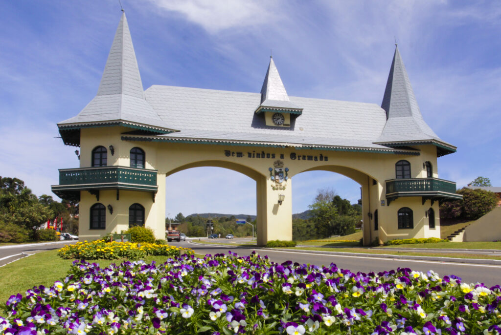 Descrição da imagem: A foto mostra, em destaque, um portal bege claro com telhado cinza claro em que lê-se ”Bem-vindo a Gramado”. Há duas torres laterais com sacadas. Em primeiro plano, há um canteiro de flores roxas. Crédito: Cleiton Thiele