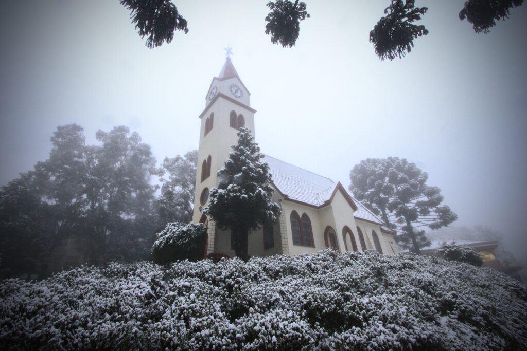 Crédito: Cleiton Thiele. [Descrição da imagem: A foto mostra o lado exterior de uma igreja com janelas e telhado vermelhos, além de uma torre com um relógio. Ao redor, há árvores e hortênsias cobertas de neve.]