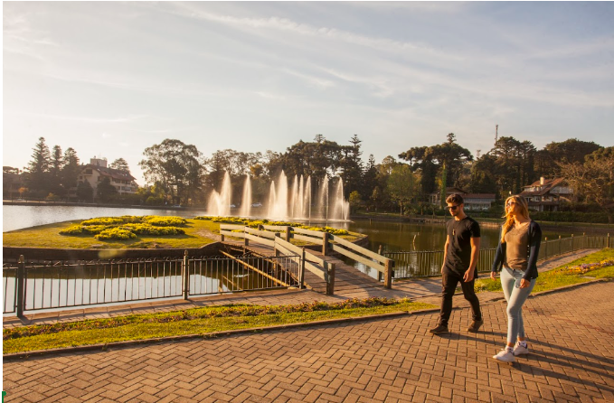 Crédito: Cleiton Thiele. [Descrição da imagem: A foto mostra uma área arborizada com um homem e uma mulher caminhando na calçada. Ao fundo, há um lago com chafariz e uma ilha com gramado ligada à calçada por uma ponte de madeira.]