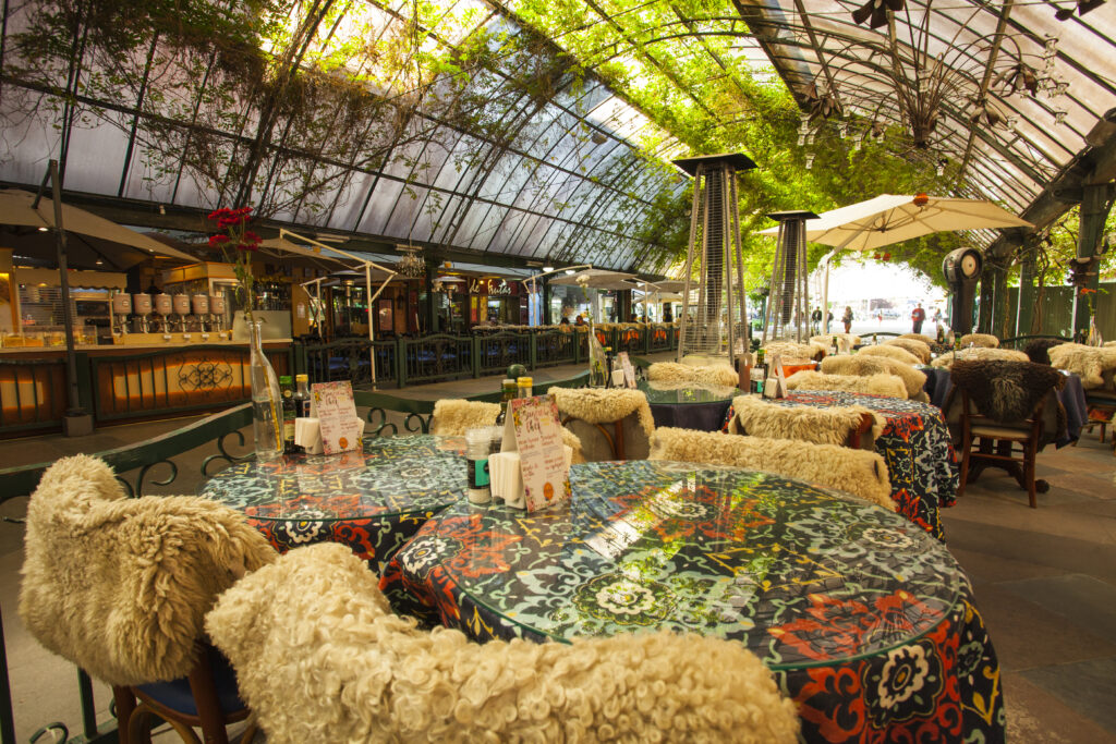 Crédito: Cleiton Thiele [Descrição da imagem: A foto mostra uma rua com uma cobertura transparente tomada por folhas verdes. Abaixo, ao nível da câmera, há várias mesas com panos coloridos, cadeiras com pelegos e aquecedores.]