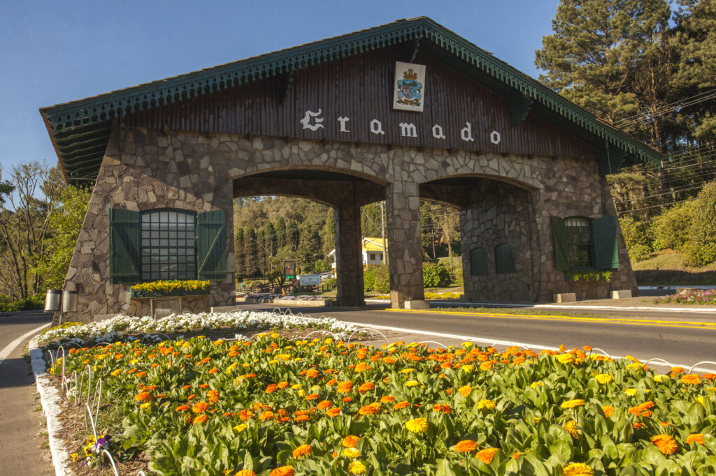Crédito: Cleiton Thiele
Descrição da imagem: A foto mostra um pórtico construído com uma base de pedra e um telhado de madeira. Há duas passagens e entre elas lê-se “Gramado”. Há uma janela verde em cada lado. No primeiro plano, aparece um canteiro de flores amarelas.
