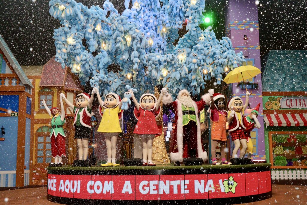 Crédito: Divulgação/Vila da Mônica Gramado [Descrição da imagem: Na foto, dois duendes, o Papai Noel e os personagens Cebolinha, Magali, Mônica, Marina e Cascão estão de mãos dadas e erguidas em um palco. Todos vestem gorros vermelhos de Natal. Neve artificial flutua ao redor deles e, ao fundo, há uma árvore com diversas pelúcias azuis do Sansão.]